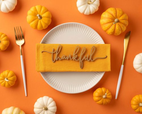 A festive Thanksgiving table setting featuring a plate, gold cutlery, and a thankful napkin surrounded by small pumpkins on an orange background